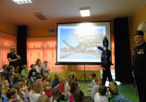Dzieci siedzą na dywanie i patrzą na projektor, na którym górnik pokazuje ilustrację.