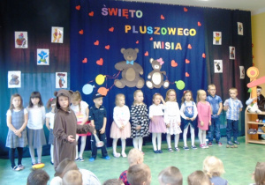 Dzieci ubrane odświetnie stoją ustawione przed kolorową dekoracją.