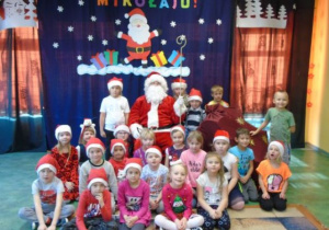 Dzieci siedzą i stoją.Razem z nimi jest Mikołaj. W tle widać kolorową dekorację.