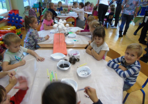 Dzieci siedzą przy stolikach. Na stolikach znajdują się pojemniki z materiałami plastycznymi.