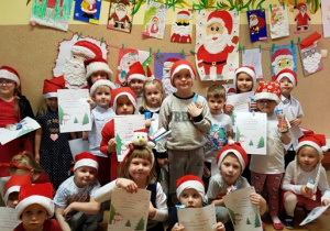 Grupa dzieci ubrana w mikołajkowe czapki.Dzieci trzymają dyplomy. Na ścianie widać powieszone na ścianie obrazki z portretami św. Mikołaja.