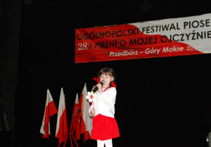 Dziewczynka ubrana w czerwoną spódniczę i białą bluzkę stoi na scenie i śpiewa.