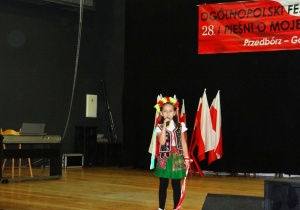 Dzieczynka ubrana w strój ludowy stoi na scenie i śpiewa.