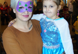 Dzieczynkę ubraną w strój księżniczki trzyma na rękach mama, która ma maskę na głowie.