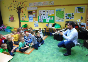 Grupa dzieci siedzi na dywanie.Muzyk z trąbką kuca przed dziećmi i pokazuje im instrument.