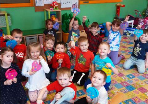 Dzieci siedzą na dywanie, trzymają w ręku kolorowe serduszka, które same wykonały.
