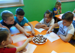 Dzieci siedza przy stole i jedzą pączki posypane cukrem pudrem.