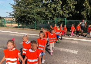 Dzieci w pomarańczowych kamizelkach odblaskowych trzymając się za ręce przechodzą przez przejście dla pieszych.