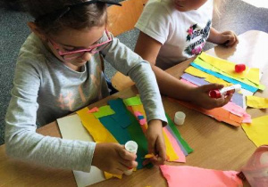 Dzieci siedzą przy stole i robią prace plastyczną wykorzystując klej i kolorowy papier.