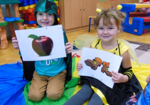 Dzieci siedza na chuście animacyjnej, Chłopiec trzyma obrazek jabłka, dziewczynka buty.