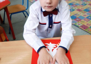 Chłopiec siedzi przy stoliku i odbija swoje dłonie na czerwonej kartce.