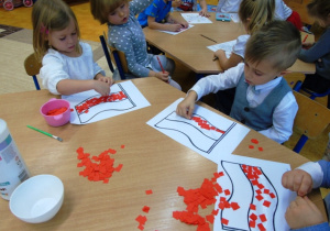 Dzieci siedzą przy stoliku i wyklejają flagi czerwonym papierem.
