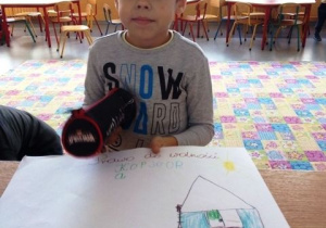 Chłopiec trzyma swoja pracę plastyczną na której jest narysowany i pokolorowany duży dom