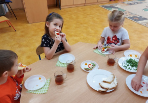 Dzieci siedza przy stolikach i jedzą kanapki.