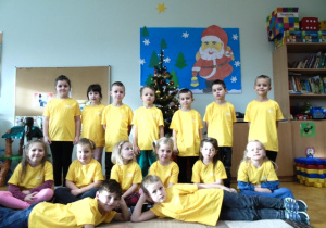 Dzieci w żółtych koszulkach stoją i siedzą na dywanie.