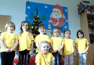 Dzieci ubrane w żółte koszulki stoją przed choinką.