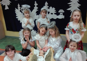 Dziewczynki w strojach aniołków siedzą obok figurki Jezuska, która leży w żłóbku.
