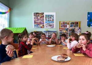 Dzieci siedzą przy stoliku i jedzą pączki.