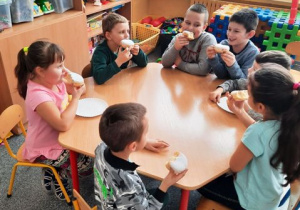 Dzieci siedzą przy stoliku i jedzą pączki.