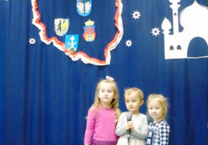 Trzy małe dziewczynki stoją na granatowym tle jednaz nich trzyma w rękach mikrofon