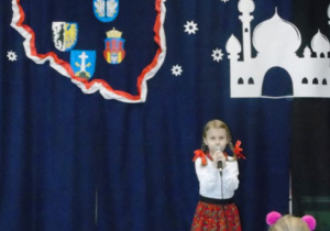 Dziewczynka w stroju góralskim stoi z miokrofonem za nią tło z konturem Polski i biały zamek z papieru