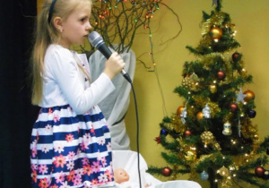 Dziewczynka odświętnie ubrana trzyma mikrofon a w tle kolorowa chinka i drzewko z lampakmi