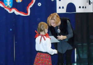 Kobieta ubrana na ciemno wręcza dyplom dziewczynce w góralskim stroju