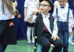 Chłopiec siedzina krześle ma na głowie kapelusz a w ręku laskę obok dziewczynka z mikrofonem w dłoni