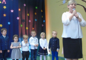Kobieta w białej bluzce i czarnej spódnicy trzyma mikrofon dzieci są w tle