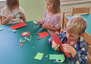 Dzieci siedzą przy stoliku i wycinają elementy z papieru.