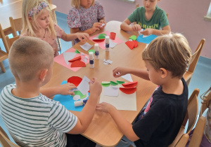 Dzieci siedzą przy stoliku i przyklejają wycięte elementy z papieru.