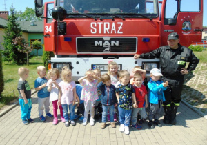 Dzieci stoją przez czerwonym samochodem przy nich strażak w czarnym stroju