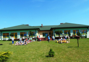 Duży zielony budynek przed nim siedzą dzieci na trawie