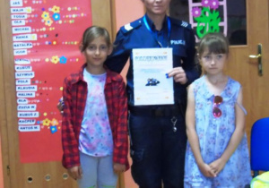 Policjantka stoi pomiędzy dwowma dziewczynkami i trzyma w ręku Podziękowanie w formie dyplomu