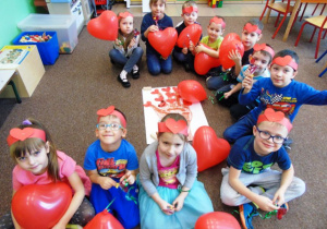 Dzieci z grupy Stokrotki siedzą na dywanie w czerwonych opaskach na głowie, trzymając w ręku czerwony balonik serce oraz lizaka